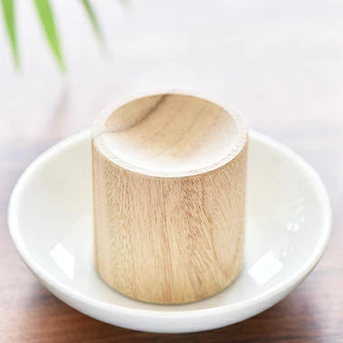 一個精緻呈圓柱狀的日本 KUSU HANDMADE 品牌天然樟木擴香座特寫照片。擴香座由紋理清晰的九州樟木雕刻而成，頂部設有微凹的圓形精油滴槽。一滴天然精油正滴落於木質凹槽中，呈現出溫潤、自然且充滿淨化能量的日系簡約工藝美感，展現出能強效除穢避邪、穩定空間氣場與催旺木行事業運的玄學氛圍。
